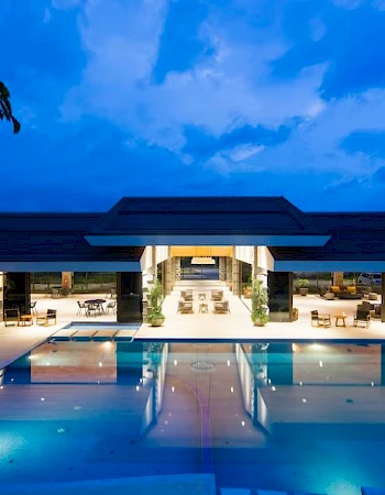 A luxurious resort with a large illuminated pool and open lounge area under a modern pavilion, set against a twilight sky.