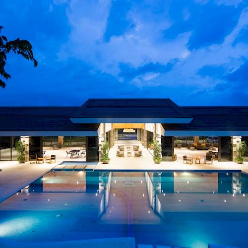 The image shows a luxurious, modern building with a large illuminated pool in front, surrounded by lounge chairs and lit up against an evening sky.