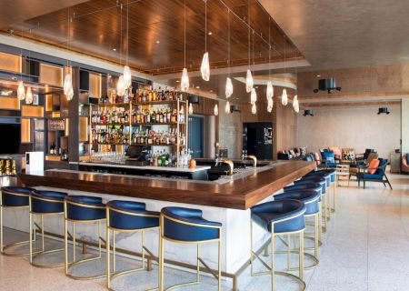 A modern bar with blue and gold stools, a well-stocked liquor shelf, pendant lights, and a seating area in the background.