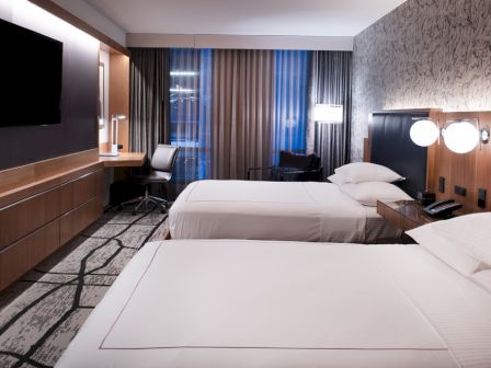 A modern hotel room with two neatly made double beds, a wall-mounted TV, a desk with a chair, and stylish lighting and decor.