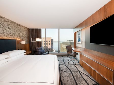 A modern hotel room features a large bed, a sleek desk with a chair, a flat-screen TV, and a window with a view of buildings and water.