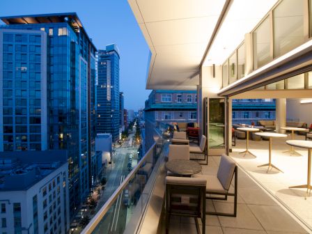 A modern rooftop patio with tables and chairs overlooks a city street lined with tall buildings, captured during evening twilight.