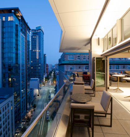 An upscale, modern balcony overlooking a city street, with outdoor seating and tables, surrounded by tall buildings lit up in the evening.