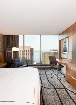 A modern hotel room with a bed, desk, chair, and large TV. The room features a large window with an urban view and stylish decor.