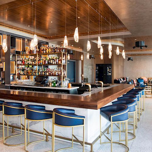 The image shows a modern bar with blue and gold chairs, pendant lights, and a well-stocked bar area in a stylish interior space.