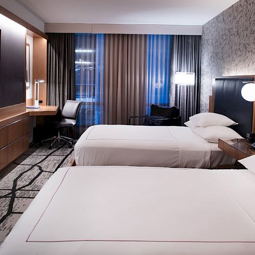 The image shows a modern hotel room with two neatly made beds, a flat-screen TV, a desk with a chair, and stylish lighting and decor on the walls.