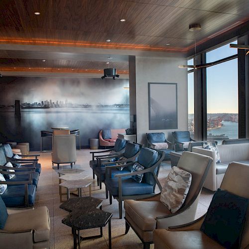 A modern lounge with multiple plush chairs, side tables, large windows offering a waterfront view, and a wall mural of a city skyline.