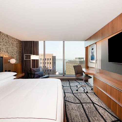 A modern hotel room features a large bed, a wall-mounted TV, a desk with a chair, and a chair by the window overlooking a city view.